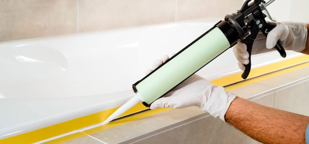 Caulk Not Sticking to Shower Tile? Diagnose It Before Recaulking Gloved hands using a caulking gun to apply a bead of sealant along a masked joint between a bathtub and tile wal