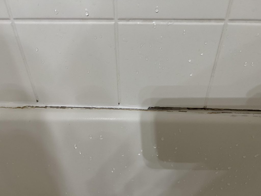 Caulk Not Sticking to Shower Tile? Diagnose It Before Recaulking Close-up of a deteriorated caulk bead along a shower tub joint, with grime and gaps where it has separated from the tile, surrounded by water droplets on the wet tile wall